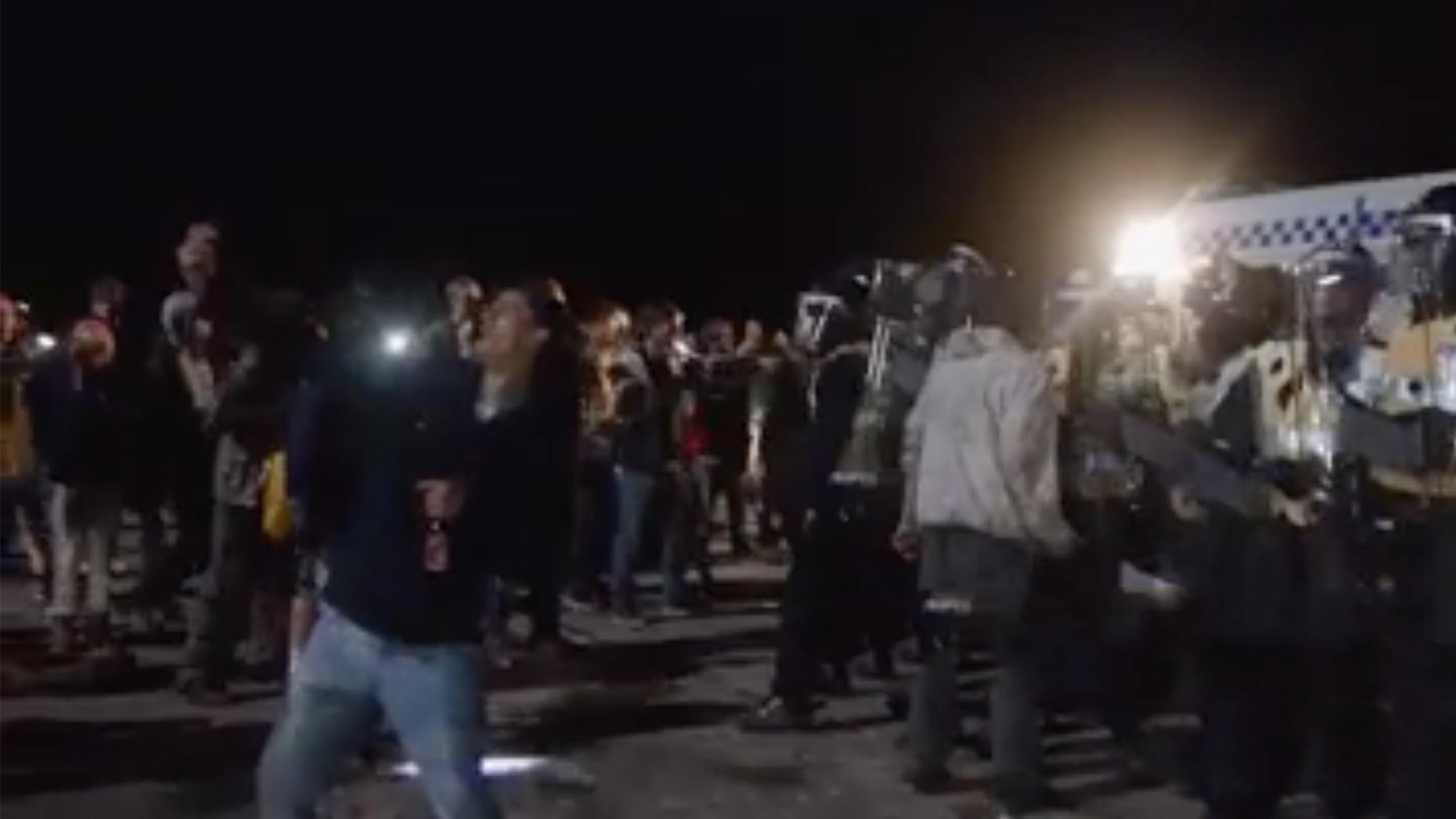 Other officers are seen holding a line of revellers back and dodging incoming missiles from the crowd, while a man is taking a selfie with the scene.  Screenshot: Chris Shaw